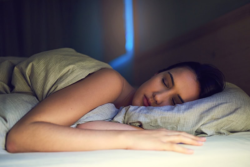 3 Health Benefits of Using Your AC While Sleeping. Photo of a young woman sleeping soundly in her bed.