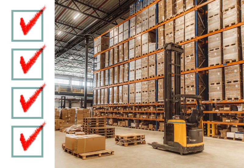 A large industrial warehouse filled with tall orange and blue shelving racks packed with stacked cardboard boxes and pallets, with a yellow and black reach truck parked in the foreground.